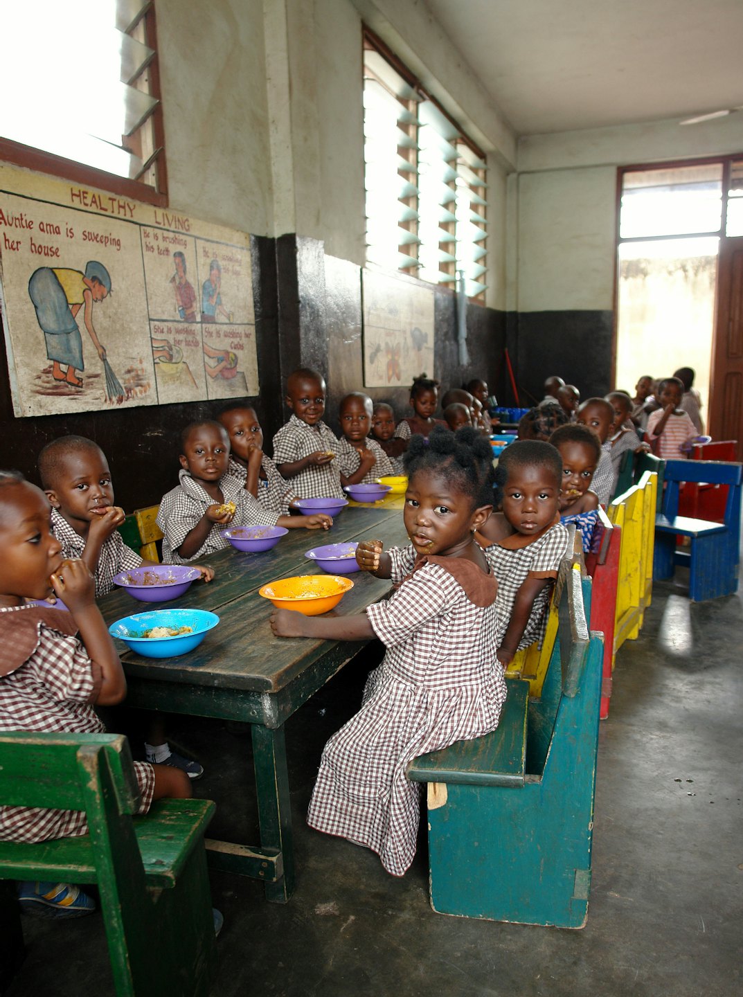Children in plaid uniforms eat lunch at tables with colorful bowls.